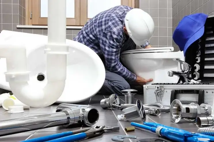 Obrero realizando trabajos de fontanería para instalar un retrete. Por el suelo se ven herramientas de fontanería y un bidet listo para ser instalado.